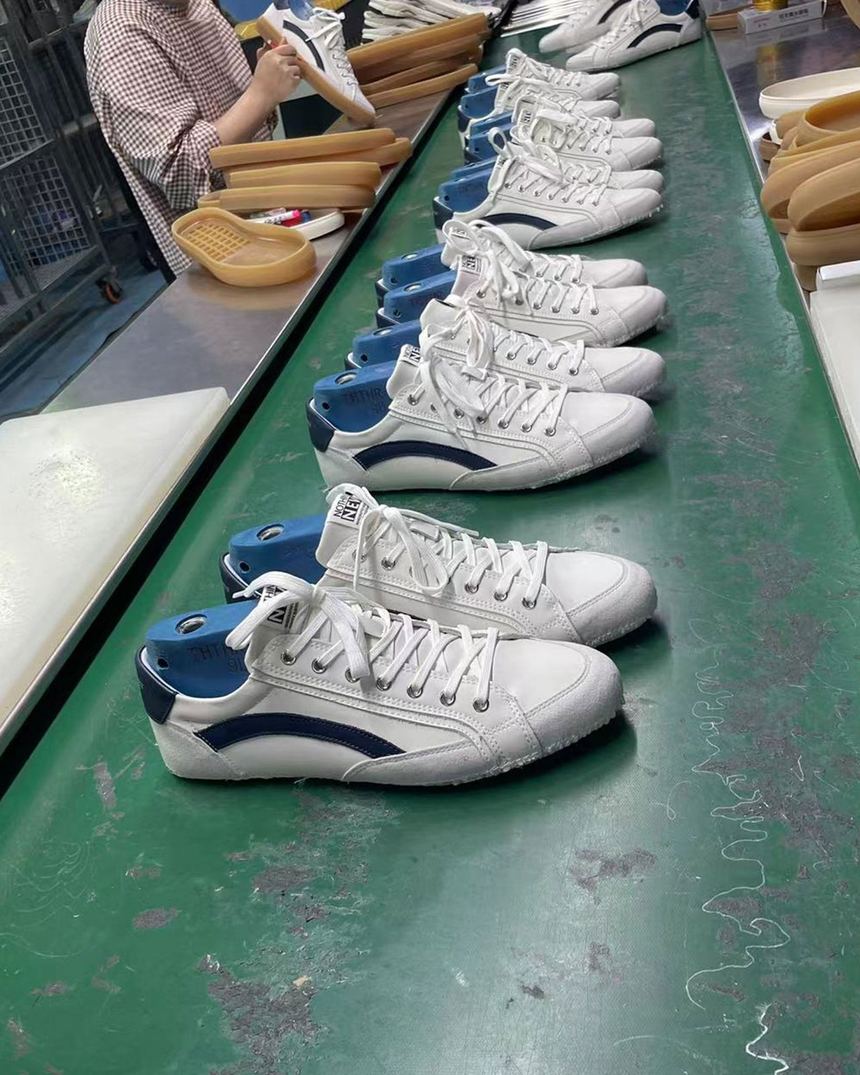 Production at one of our partner factories: white and blue sneakers lined up on a green conveyor, with soles and a worker's hands at the edge of the line.